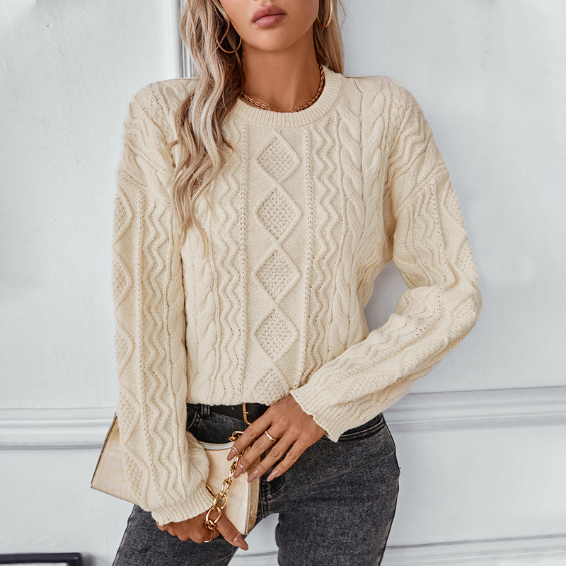 Une femme porte un pull blanc à torsades, orné d'un motif losanges et de torsades verticales. Ce pull à la coupe droite possède des manches longues et des bords-côtes au col, aux poignets et à la base. Elle l'associe à un jean noir et un petit sac à main beige à chaîne dorée, pour un look élégant et moderne. La douce lumière met en valeur la texture du tricot et la teinte chaude du pull.