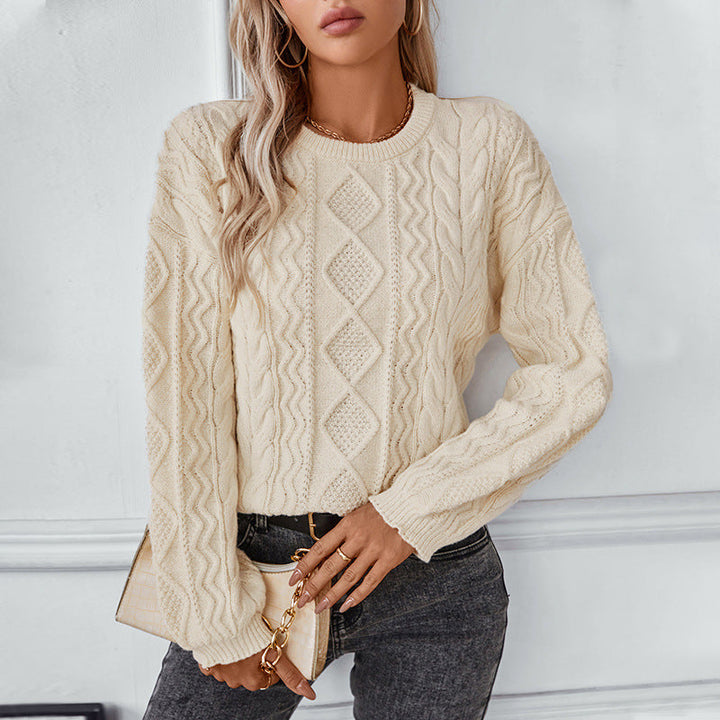 Une femme porte un pull blanc à torsades, orné d'un motif losanges et de torsades verticales. Ce pull à la coupe droite possède des manches longues et des bords-côtes au col, aux poignets et à la base. Elle l'associe à un jean noir et un petit sac à main beige à chaîne dorée, pour un look élégant et moderne. La douce lumière met en valeur la texture du tricot et la teinte chaude du pull.