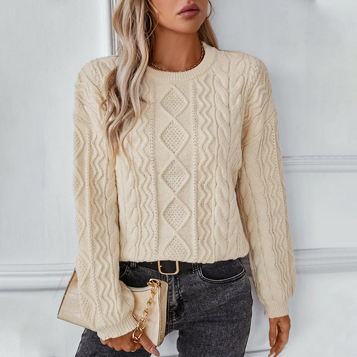 Une femme porte un pull blanc en maille torsadée au design raffiné, orné de motifs losanges et de torsades verticales. Ce pull, à la texture douce et épaisse, est idéal pour les journées fraîches. Ses manches longues légèrement bouffantes et ses bords-côtes lui confèrent une touche de confort et d'élégance. Elle complète sa tenue avec un jean foncé et un sac à main beige à chaîne dorée, pour un look moderne et chic dans un intérieur lumineux.