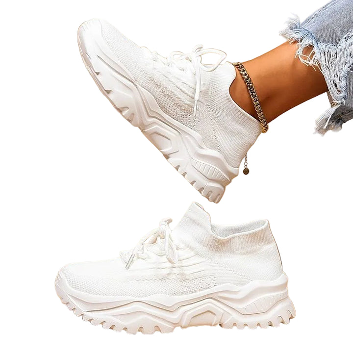 Baskets blanches en maille respirante avec lacets, semelle épaisse crantée ultra confortable, portées avec un jean clair sur fond beige.