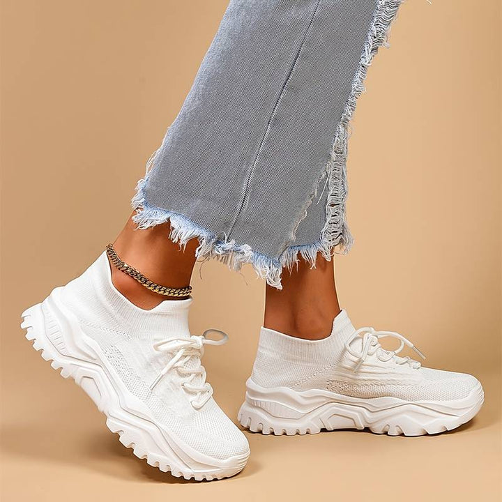 Baskets blanches pour femme en maille avec lacets, semelle épaisse crantée, vue de profil portées avec un jean gris effiloché et une chaîne de cheville, sur fond beige.