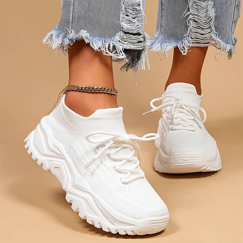 Baskets blanches pour femme en maille avec lacets, semelle épaisse crantée, portées de face avec un jean effiloché et une chaîne de cheville, sur fond beige.