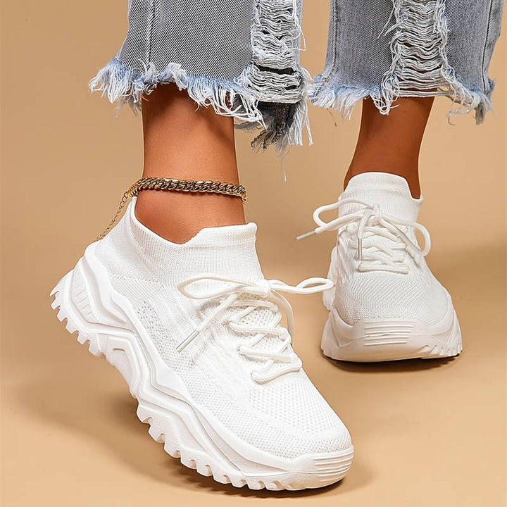 Baskets blanches pour femme en maille avec lacets, semelle épaisse crantée, portées de face avec un jean effiloché et une chaîne de cheville, sur fond beige.