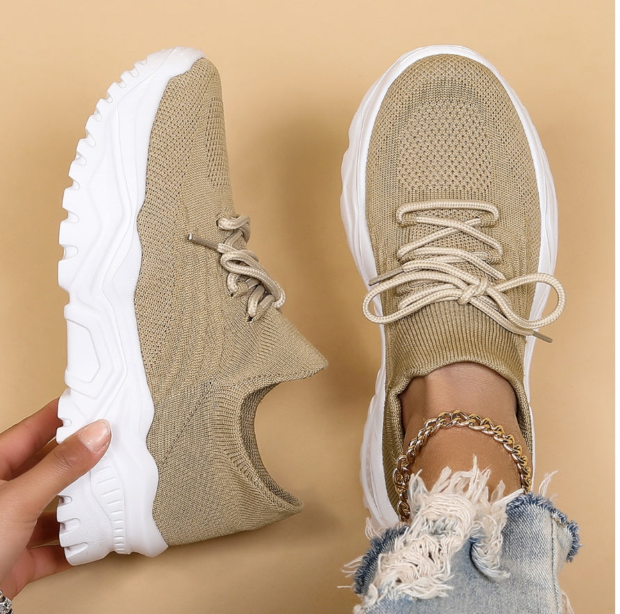 Baskets beiges pour femme en maille avec lacets, semelle épaisse crantée blanche, une chaussure portée et l’autre tenue à la main, avec jean effiloché et chaîne de cheville, sur fond beige.