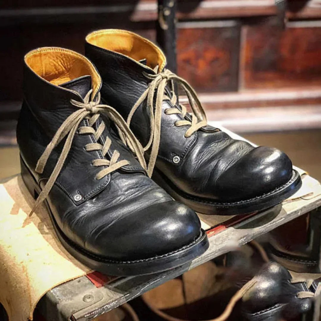 Une paire de bottes en cuir noir pour homme posée sur une surface en bois, avec des lacets beiges et une doublure intérieure marron clair. Les chaussures affichent un style vintage et robuste, mettant en valeur leur finition soignée et leur design classique. Idéales pour une allure élégante et durable au quotidien.