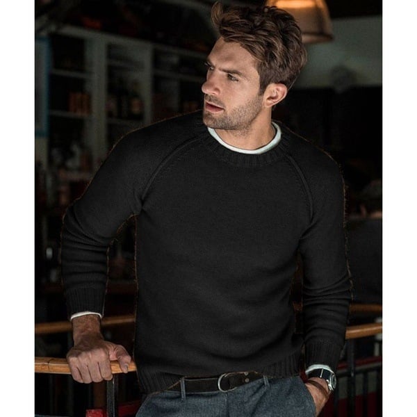 Un homme portant un pull noir en tricot avec un col rond. Il a un look moderne et élégant, avec des cheveux courts et une posture détendue, vêtu d'un pantalon gris et d'une ceinture noire, posé dans un cadre intérieur chaleureux.