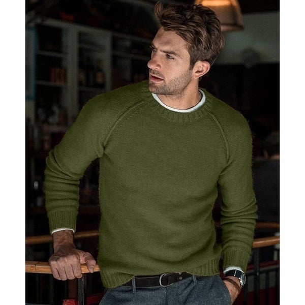 Un homme portant un pull tricoté vert olive avec un col rond. Il affiche un look moderne et élégant, avec des cheveux courts et une posture détendue, vêtu de pantalons gris et d'une ceinture noire, dans un cadre intérieur confortable.
