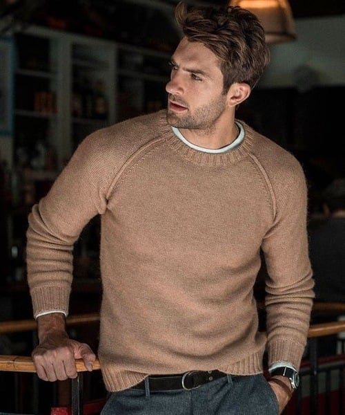 Un homme portant un pull tricoté beige avec un col rond. Il adopte une posture détendue avec une main appuyée sur une barrière en bois, vêtu de pantalons gris et d'une ceinture noire, affichant un style moderne et élégant dans un environnement intérieur chaleureux.