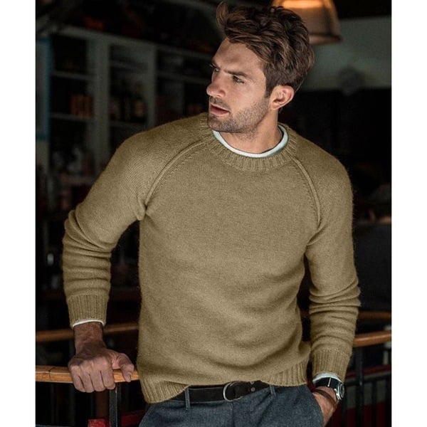 Un homme portant un pull tricoté beige avec un col rond, affichant une posture décontractée. Il a les bras croisés et porte un pantalon gris avec une ceinture noire, dans un environnement intérieur cosy et élégant.