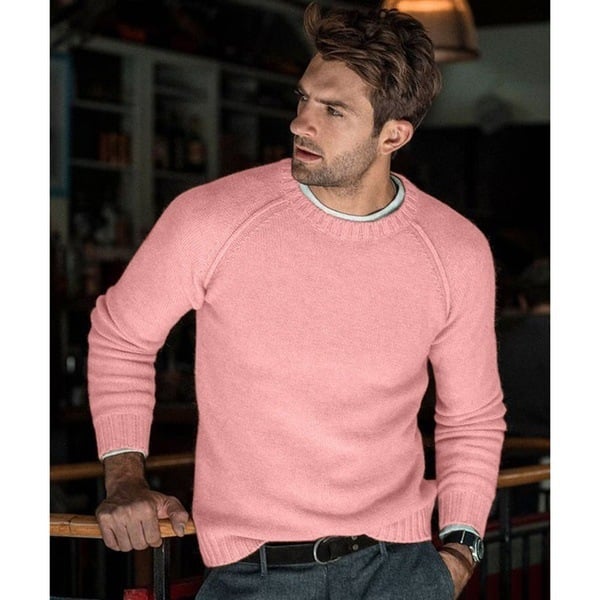 Un homme portant un pull tricoté rose avec un col rond, affichant une posture décontractée tout en se tenant dans un environnement intérieur chaleureux. Il porte un pantalon gris et une ceinture noire, avec une légère expression réfléchie sur son visage.