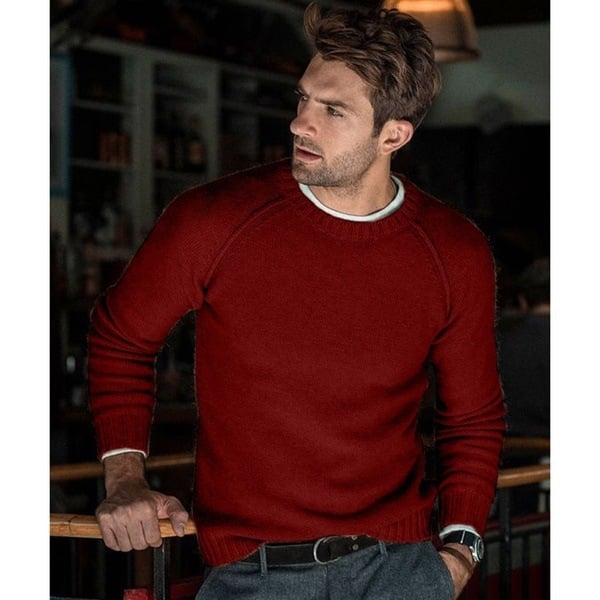 Un homme portant un pull tricoté rouge avec un col rond, dans une ambiance intérieure chaleureuse. Il a une posture détendue, se tenant d'une main sur une barre tout en affichant une expression légèrement pensive. Il porte un pantalon gris et une ceinture noire.