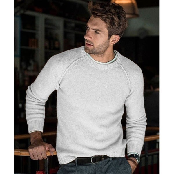 Un homme portant un pull tricoté blanc avec un col rond. Il se tient d'une main sur une barre dans un environnement intérieur avec une ambiance chaleureuse. Il porte un pantalon gris et une ceinture noire, et affiche une expression pensive en regardant sur le côté.
