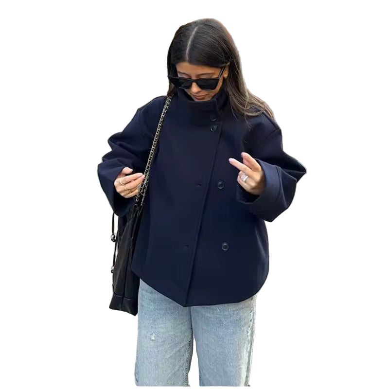 Femme portant un manteau court bleu marine à double boutonnage, associée à un jean clair et un sac à bandoulière noir. Le manteau présente une coupe ample et un col montant pour un style moderne et confortable. La scène de rue urbaine souligne une allure élégante et décontractée, parfaite pour la mi-saison.