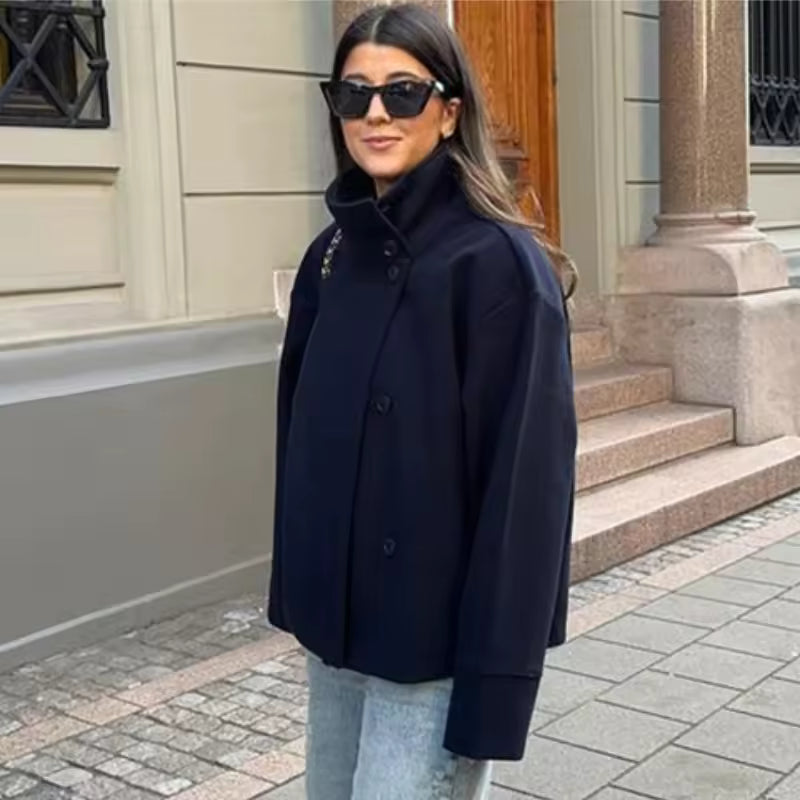 Femme élégante portant un manteau court bleu marine à double boutonnage, associée à un jean clair et des lunettes de soleil noires. La coupe ample du manteau et son col montant offrent un style moderne et raffiné, idéal pour la mi-saison. La scène urbaine en arrière-plan met en valeur une allure chic et décontractée.