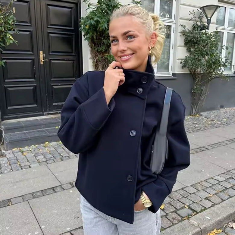 Femme élégante portant un manteau court bleu marine à double boutonnage, assorti à un pantalon clair. Le manteau présente une coupe structurée avec un col montant et de larges manches, offrant une allure moderne et sophistiquée. L’ensemble est complété par un sac à bandoulière foncé et des bijoux dorés, idéal pour un style urbain chic et tendance.
