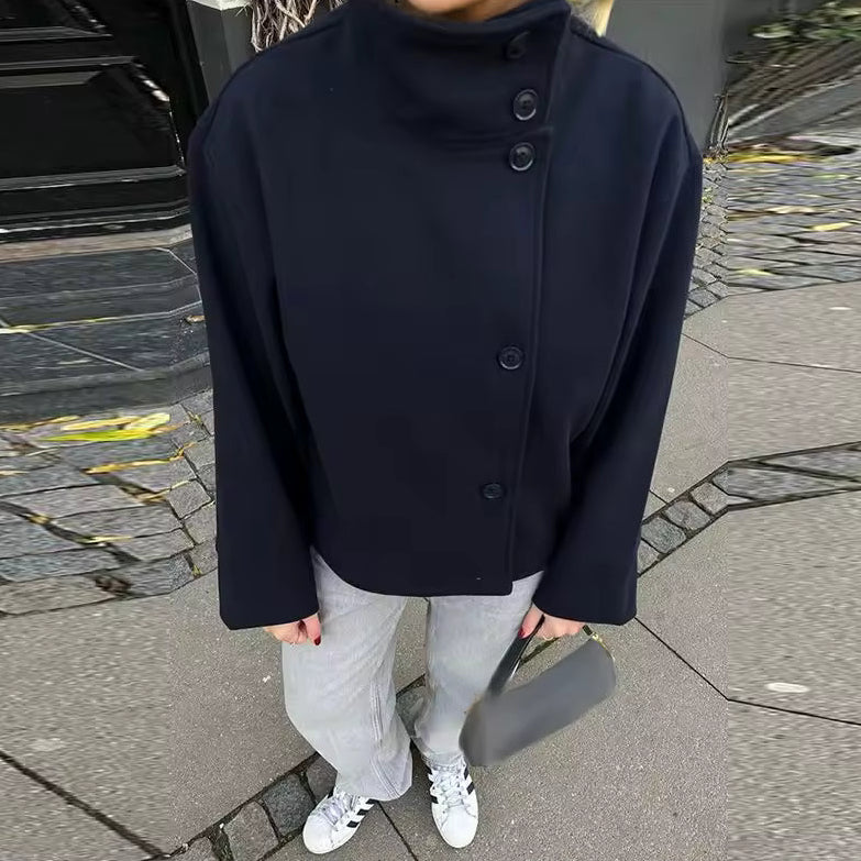 Femme portant un manteau court bleu marine à double rangée de boutons sur le côté, combiné avec un pantalon clair et des baskets blanches. Le manteau présente un col montant élégant et une coupe structurée, idéale pour un style moderne et minimaliste. L’ensemble reflète une allure chic et décontractée parfaite pour les journées fraîches en ville.