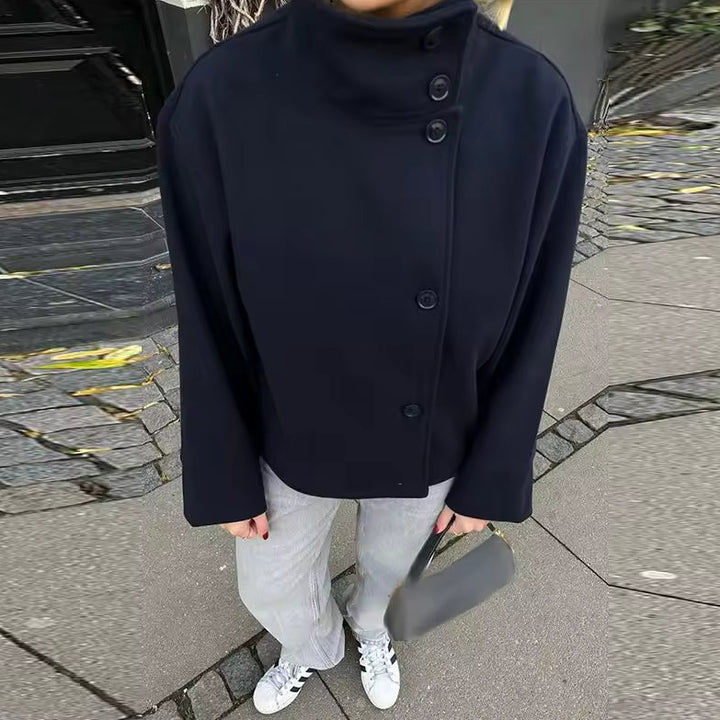Femme portant un manteau court bleu marine à double rangée de boutons sur le côté, combiné avec un pantalon clair et des baskets blanches. Le manteau présente un col montant élégant et une coupe structurée, idéale pour un style moderne et minimaliste. L’ensemble reflète une allure chic et décontractée parfaite pour les journées fraîches en ville.