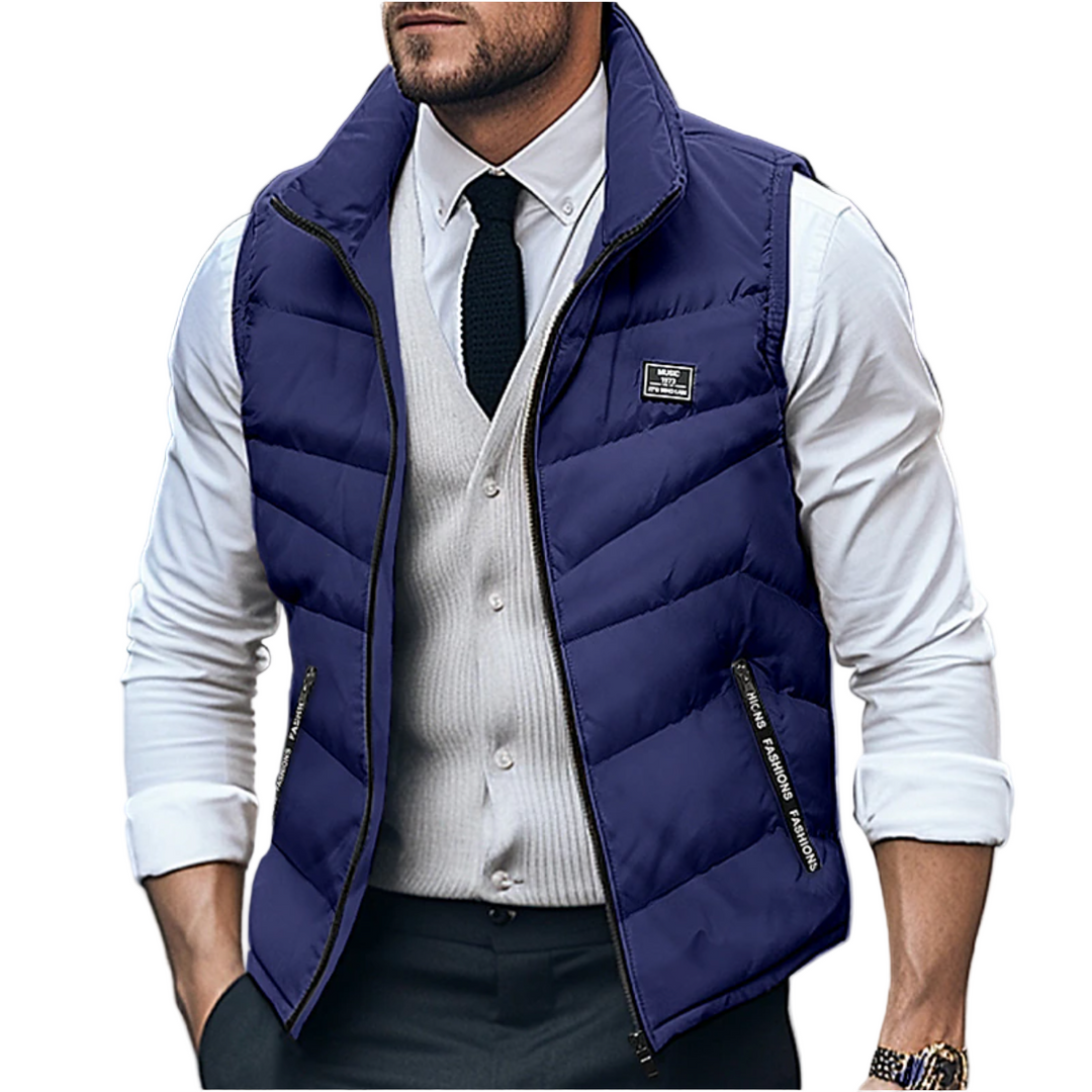 Homme portant un gilet matelassé sans manches bleu marine avec fermeture éclair, associé à une chemise blanche, un gilet beige clair et une cravate noire. Le look élégant et moderne met en valeur une tenue à la fois décontractée et chic, idéale pour les journées fraîches d’hiver en milieu urbain.