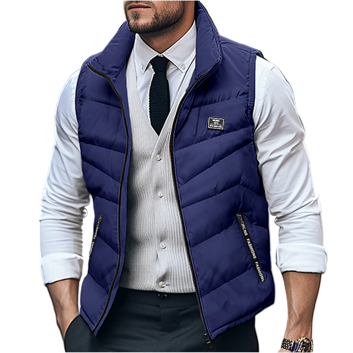 Homme portant un gilet matelassé sans manches bleu marine avec fermeture éclair, associé à une chemise blanche, un gilet beige clair et une cravate noire. Le look élégant et moderne met en valeur une tenue à la fois décontractée et chic, idéale pour les journées fraîches d’hiver en milieu urbain.