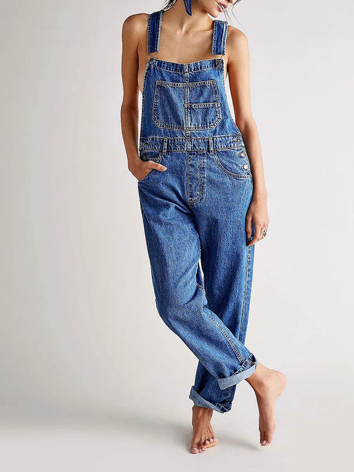 Femme portant la combinaison moderne Veloria en jean bleu, style salopette à bretelles avec poche poitrine et poches latérales, coupe ample et bas retroussés.