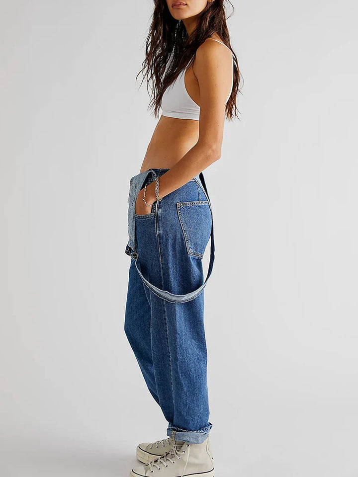 Vue de profil de la combinaison moderne Veloria en jean bleu, style salopette ample portée avec une bretelle abaissée, jambes droites retroussées et baskets.