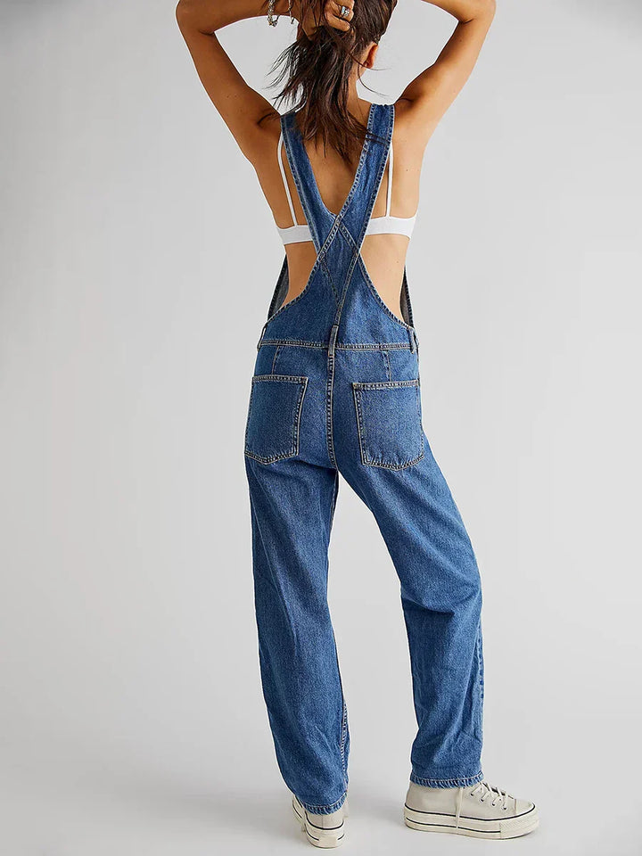 Vue de dos de la combinaison moderne Veloria en jean bleu, bretelles croisées, coupe ample avec poches arrière, portée avec brassière blanche et baskets.