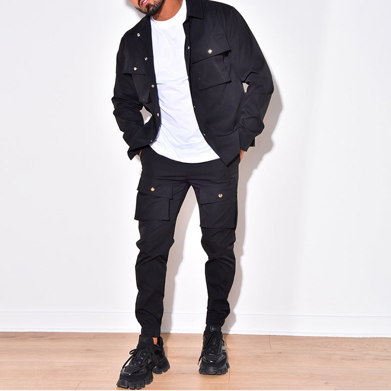 Un homme porte un ensemble de style cargo noir avec une chemise à boutons et un pantalon assorti, chacun orné de poches pratiques. Il complète son look avec des baskets noires et un t-shirt blanc. Il se tient dans une position décontractée, montrant un style urbain et moderne, sur fond neutre.