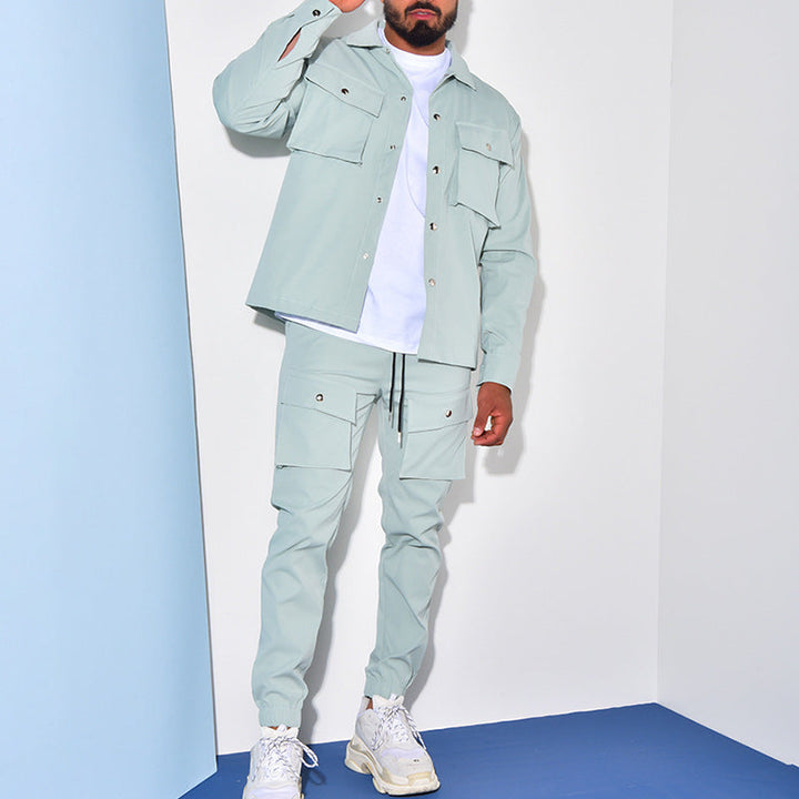 Un homme porte un ensemble cargo bleu clair, composé d'une chemise à boutons et d'un pantalon assorti. Il complète son look avec des baskets blanches et un t-shirt blanc. Il se tient dans un environnement intérieur moderne, avec des murs blancs et des éléments de design minimaliste autour de lui.