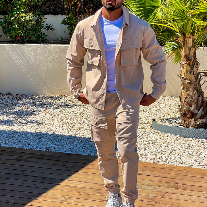 Un homme porte un ensemble cargo beige avec une chemise à boutons et un pantalon assorti, complété par des baskets blanches et un t-shirt blanc. Il marche sur un sol en bois, entouré de végétation, dans un cadre extérieur moderne et lumineux.