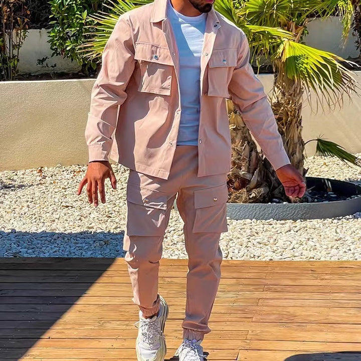 Un homme porte un ensemble cargo beige, comprenant une chemise à boutons et un pantalon cargo avec des poches. Il marche sur un sol en bois près d'un jardin avec des palmiers. Il porte des baskets blanches et un t-shirt blanc sous la chemise.