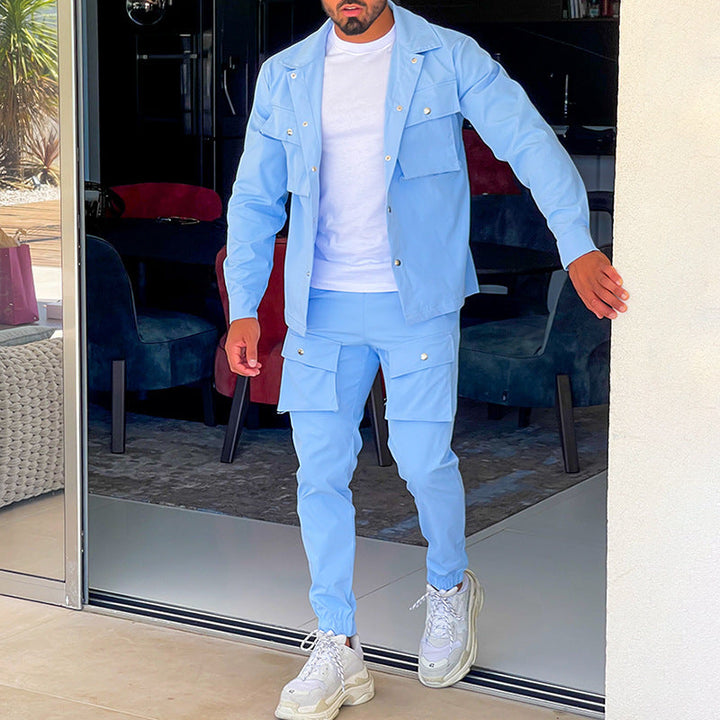 Un homme porte un ensemble cargo bleu clair composé d'une chemise à boutons et d'un pantalon cargo avec des poches. Il marche près d'une porte vitrée, avec un intérieur moderne et des sièges colorés visibles en arrière-plan. Il porte des baskets blanches et un t-shirt blanc sous la chemise.