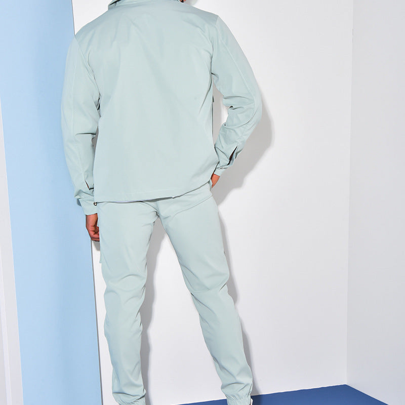 Un homme est vu de dos, portant un ensemble cargo vert menthe composé d'une chemise à manches longues et d'un pantalon cargo. Le vêtement est conçu avec des poches et des détails ajustés au niveau des poignets et des chevilles. L'homme se tient dans un environnement minimaliste avec des murs blancs et bleus derrière lui.