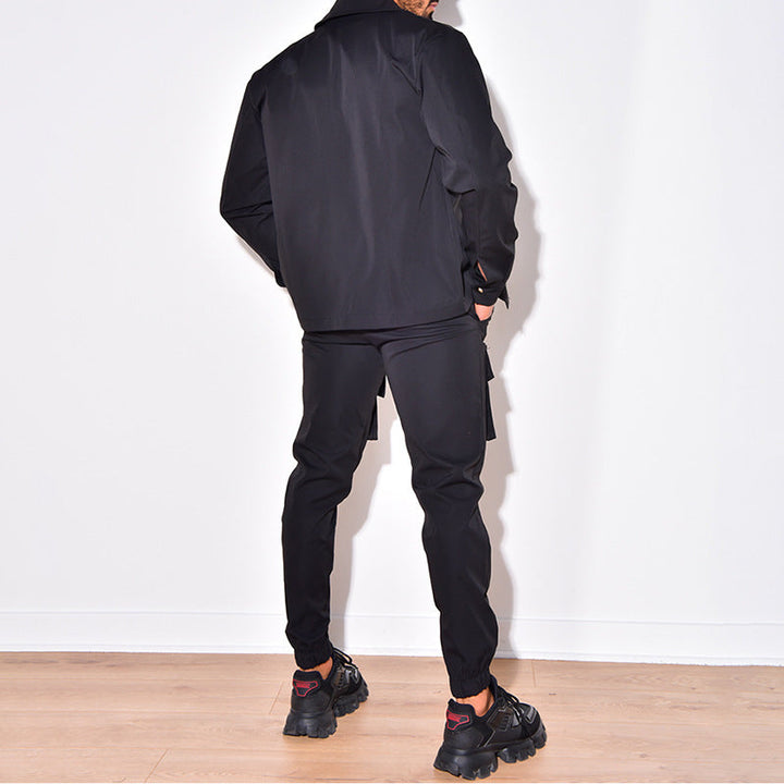 Un homme est vu de dos portant un ensemble cargo noir, composé d'une veste à manches longues et d'un pantalon cargo. Il est équipé de baskets noires et se tient dans un environnement minimaliste avec des murs blancs derrière lui.