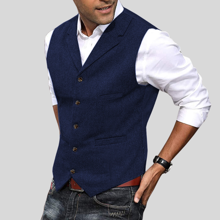 Gilet court pour hommes Virgile moderne, bleu marine à motif chevrons, coupe ajustée avec boutons et poches passepoilées, porté sur chemise blanche avec jean.