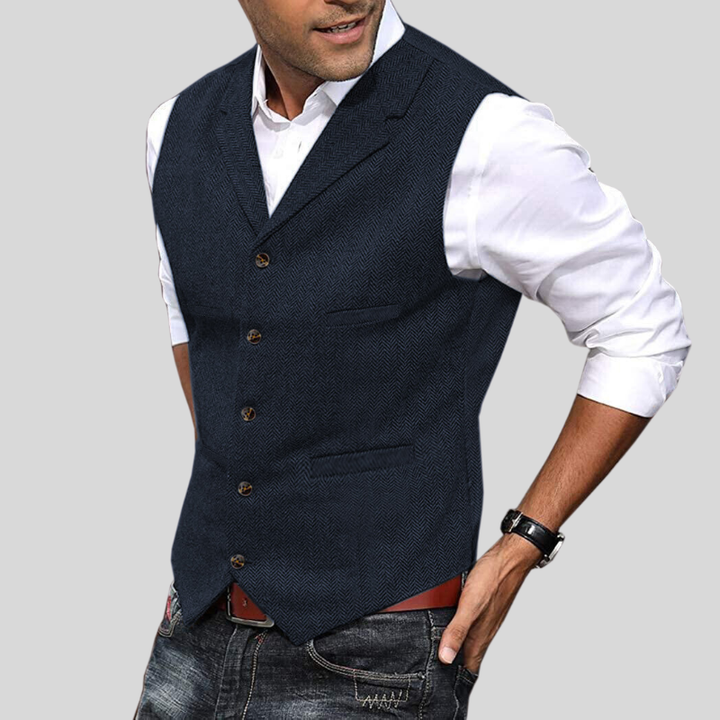Gilet court pour hommes Virgile moderne, bleu nuit à motif chevrons, coupe ajustée avec boutons et poches passepoilées, porté sur chemise blanche avec jean.
