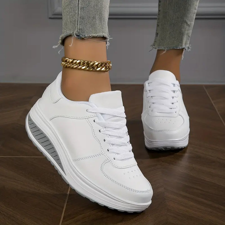Chaussures plates pour femme Wanda tendance, baskets basses noires à lacets avec empiècement blanc au talon et semelle épaisse confortable, portées avec un jean sur sol en bois.