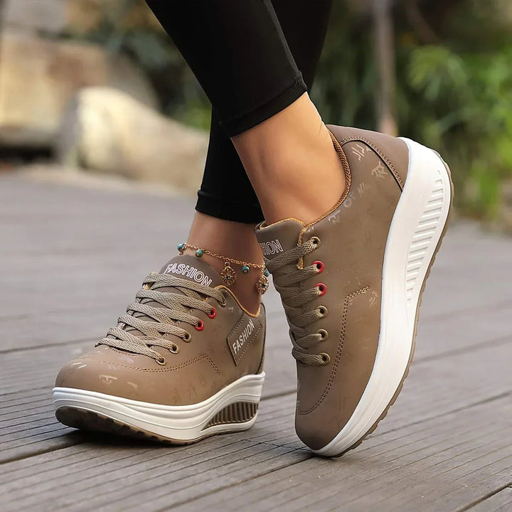 Chaussures plates pour femme Wanda tendance, baskets basses taupe à lacets avec détails et œillets rouges, semelle épaisse blanche, portées en extérieur.