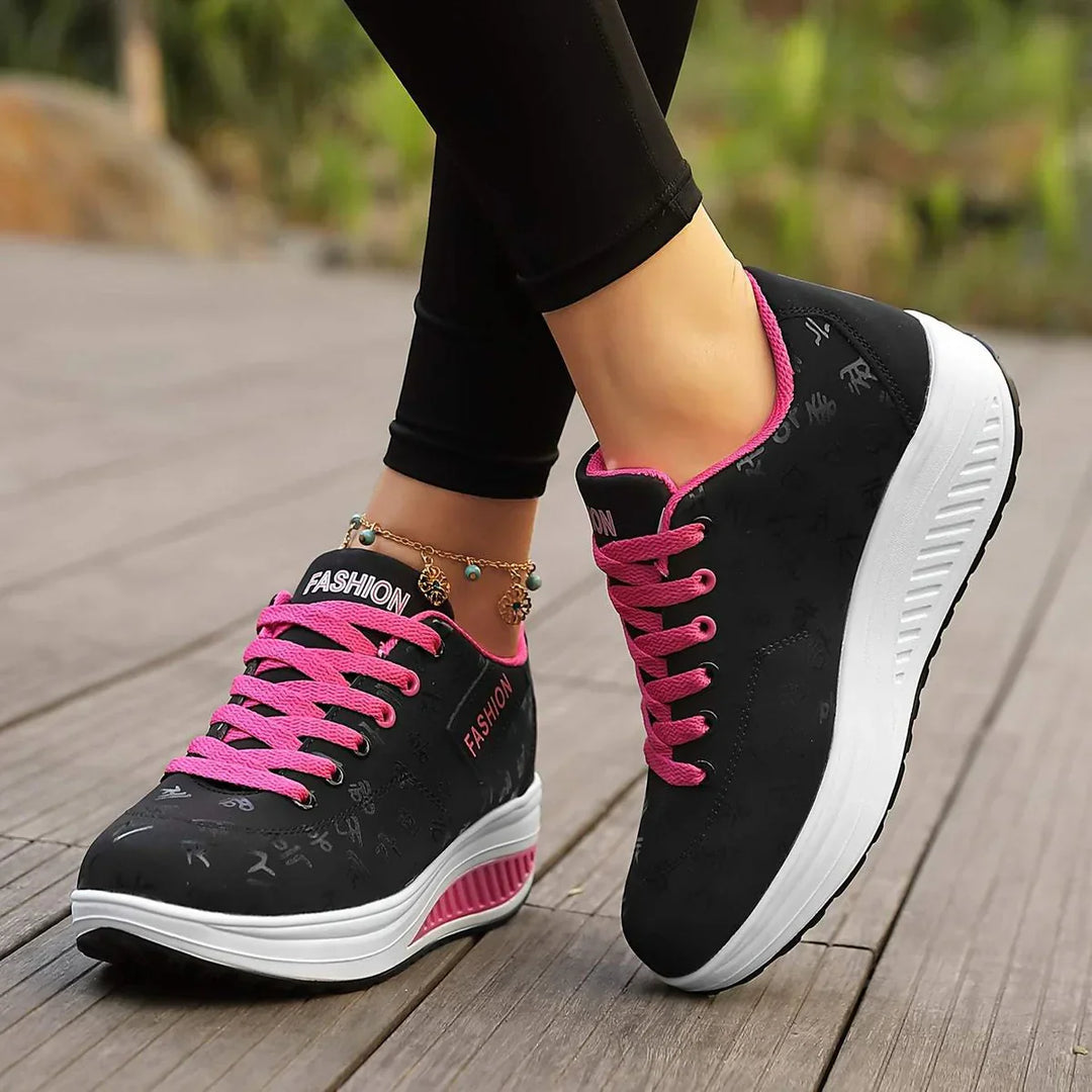 Chaussures plates pour femme Wanda tendance, baskets basses noires avec lacets roses, détails  et semelle épaisse blanche confortable, portées en extérieur sur terrasse en bois.