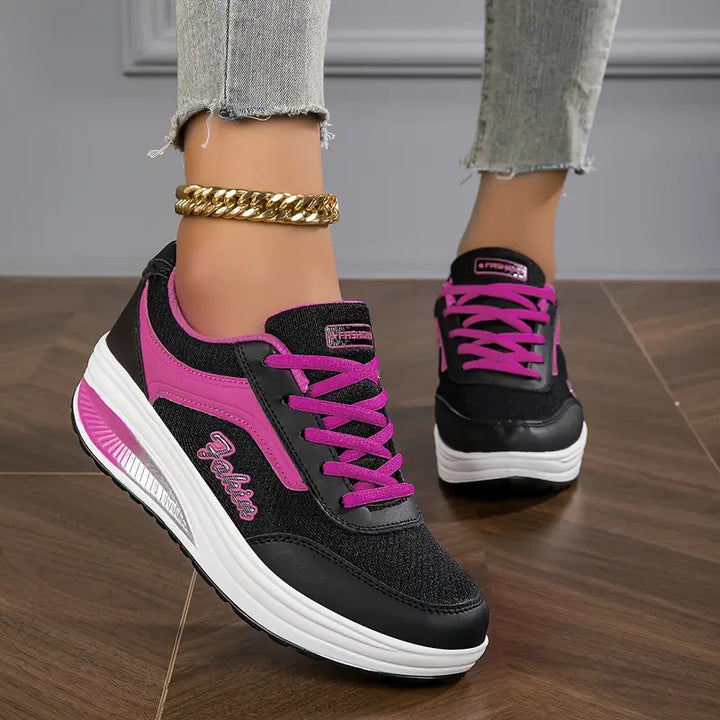 Chaussures plates pour femme Wanda tendance, baskets basses noires en mesh avec détails et lacets rose fuchsia, semelle épaisse blanche confortable, portées avec un jean sur sol en bois.