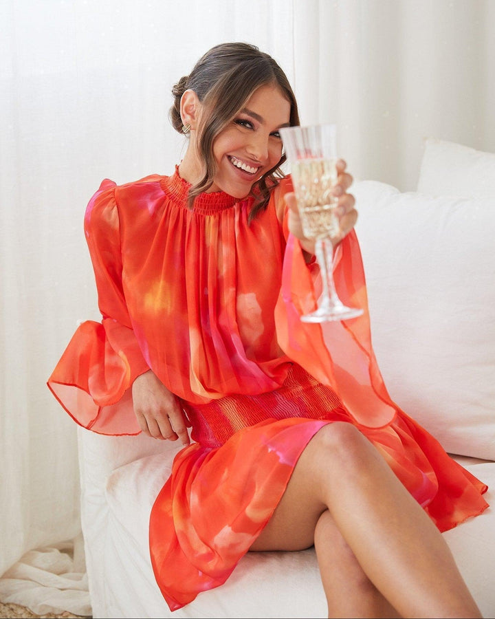 Robe mini d'été à manches longues pour femme, coloris dégradé rose et orange, avec un buste smocké et des manches évasées. Le modèle est porté par une femme souriante, tenant un verre de champagne, incarnant un style chic et festif, parfait pour les occasions estivales.