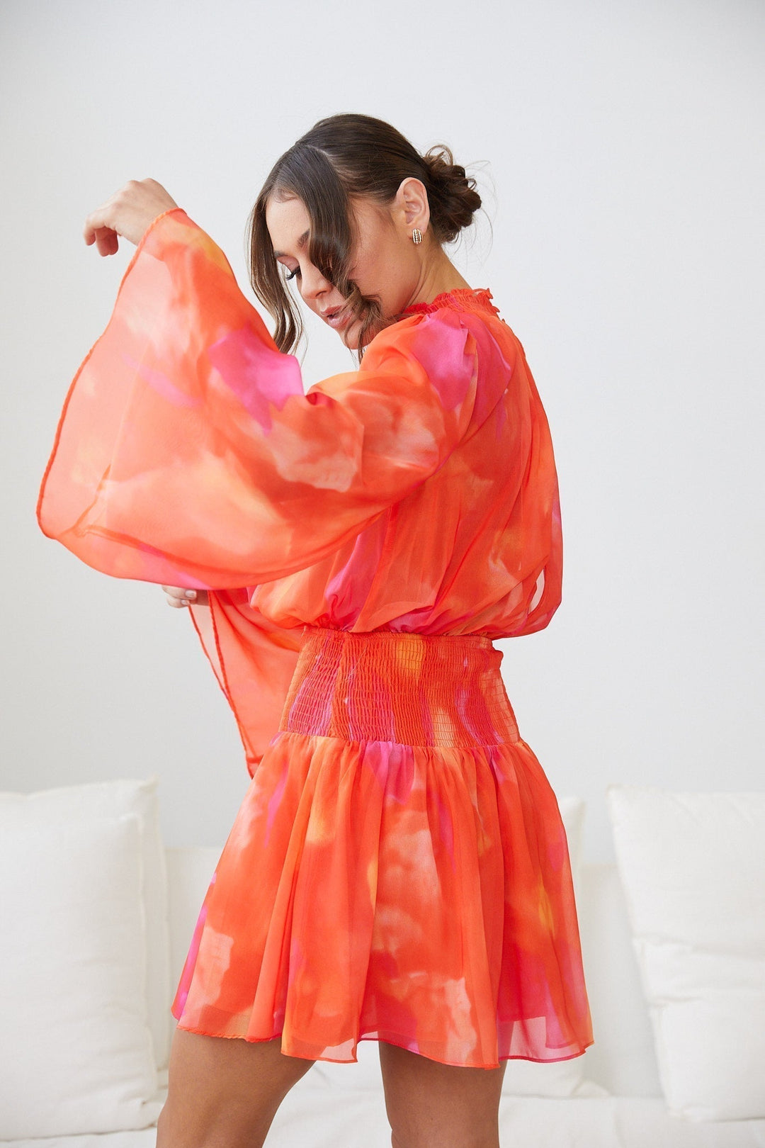 Robe mini d'été à manches longues pour femme, de style moderne avec un dégradé de couleurs rose et orange. Le modèle présente un buste smocké et des manches évasées. La femme porte la robe en la tournant avec grâce, créant une silhouette fluide et élégante, idéale pour un look estival.