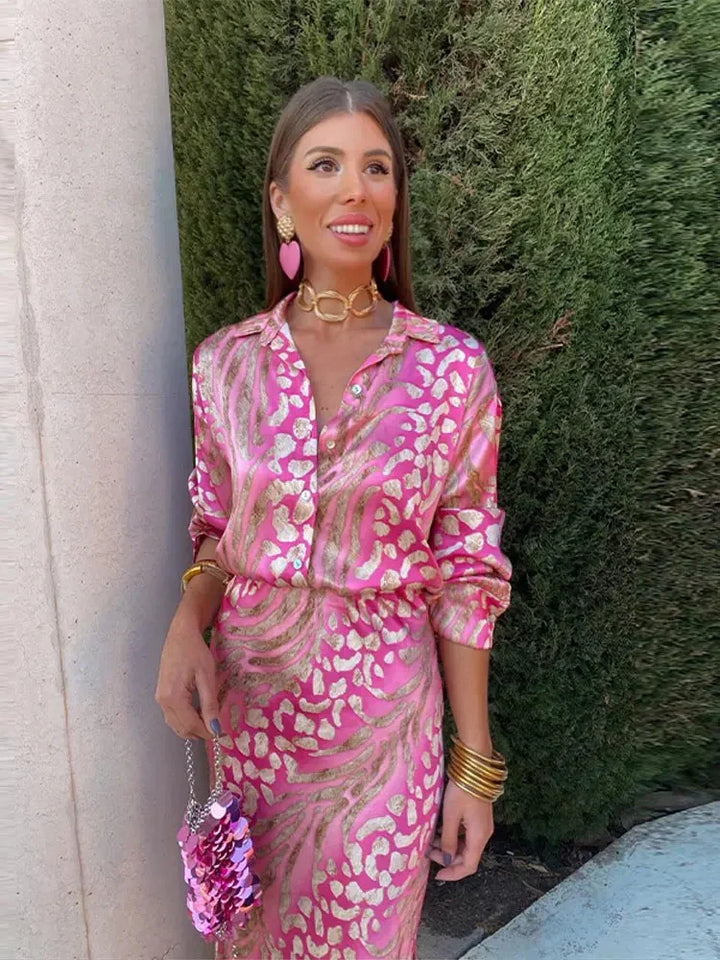 Femme portant la robe d’été Wynéa à manches longues, coupe chemise en satin rose avec imprimé doré, ceinturée à la taille, look élégant avec bijoux et pochette.