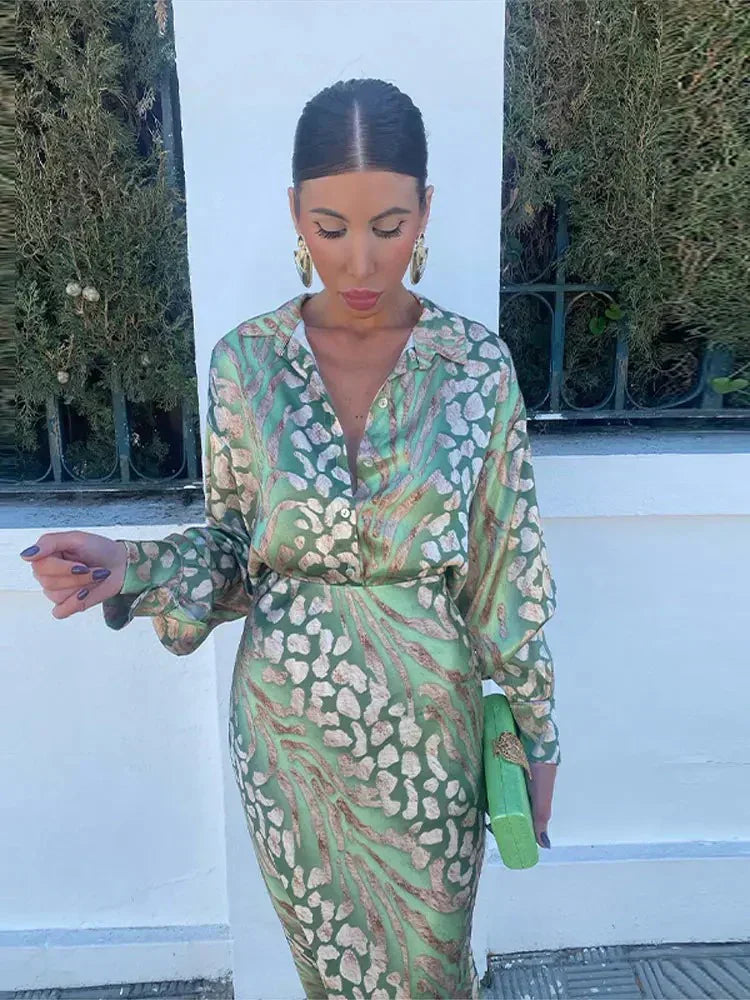 Femme portant la robe d’été Wynéa à manches longues, coupe chemise en satin vert avec imprimé doré, ceinturée à la taille, look chic avec pochette assortie.