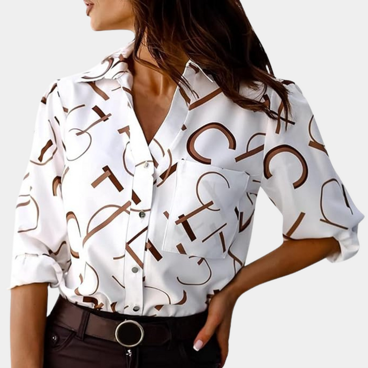 Chemise femme Xavine blanche à lettres graphiques marron portée avec un pantalon foncé. Modèle avec col classique, poche poitrine et manches retroussées, idéal pour un look tendance, moderne et élégant.