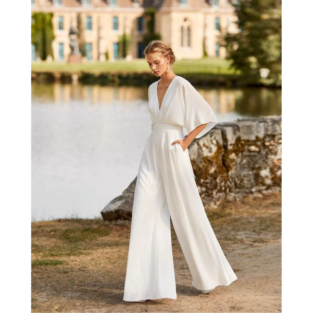 Combinaison blanche fluide à col V et manches amples, taille marquée, jambes larges, portée par un mannequin au bord d’un lac.