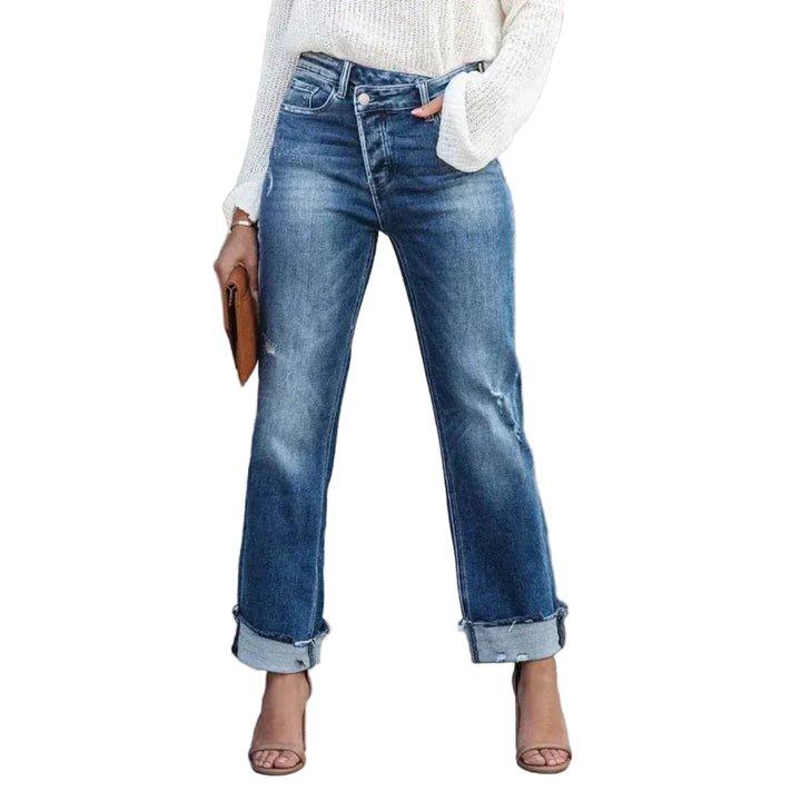 Femme portant le pantalon droit Zephyra, jean bleu taille haute à coupe droite avec bas retroussés, associé à un pull blanc et des sandales à talons  style moderne et élégant.