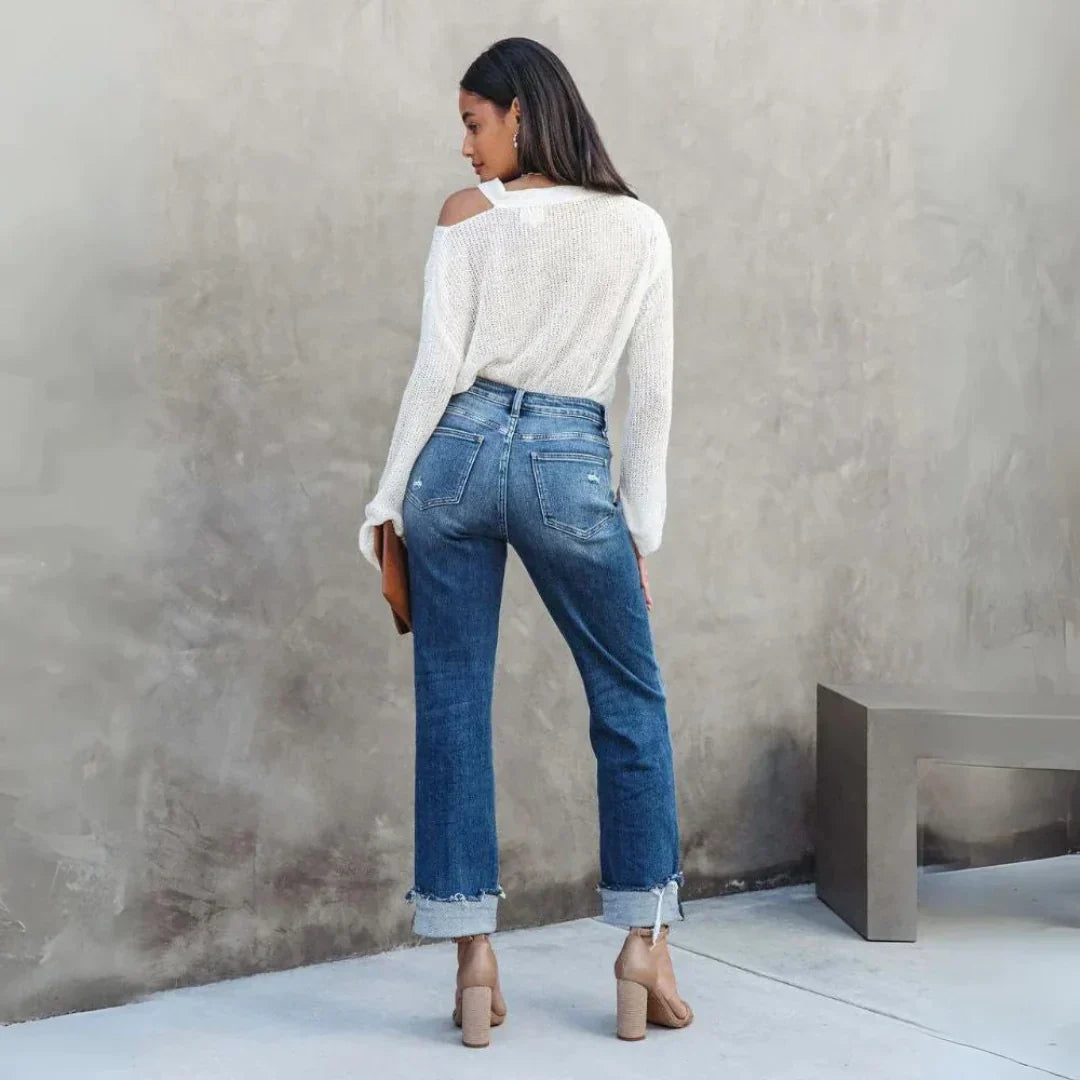 Vue de dos d’une femme portant le pantalon droit Zephyra, jean bleu taille haute à coupe droite avec bas retroussés et poches arrière, porté avec un pull blanc et des sandales à talons  look moderne et tendance.