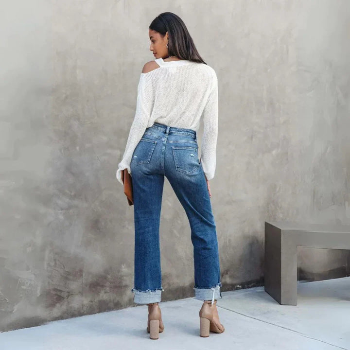 Vue de dos d’une femme portant le pantalon droit Zephyra, jean bleu taille haute à coupe droite avec bas retroussés et poches arrière, porté avec un pull blanc et des sandales à talons  look moderne et tendance.