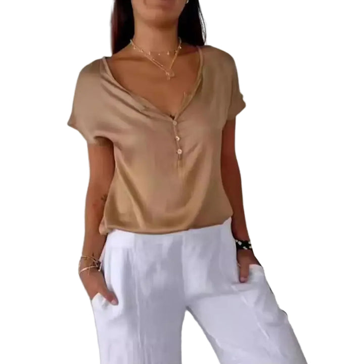 Haut léger beige à manches courtes avec un décolleté en V et des boutons délicats. Il est porté avec un pantalon blanc ample, créant un look moderne et décontracté, parfait pour les journées estivales.