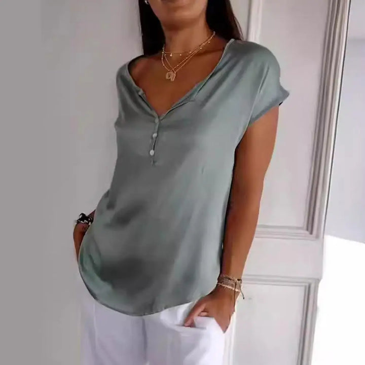 Haut à manches courtes de couleur vert olive avec un décolleté en V et des boutons délicats. Ce look moderne et décontracté est porté avec un pantalon blanc ample, créant une tenue élégante et confortable, idéale pour les journées ensoleillées.